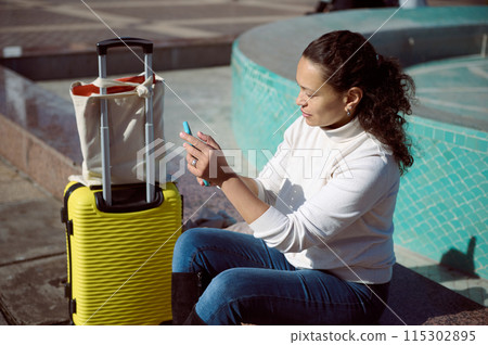 Woman traveling with yellow suitcase and using smartphone outdoors in sunny setting Woman traveling with yellow suitcase and using smartphone outdoors in sunny setting 115302895