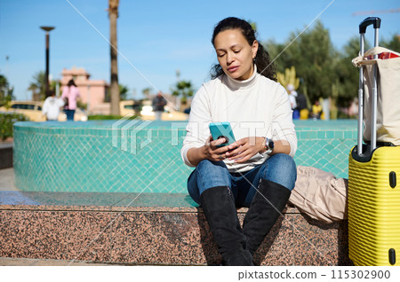 Woman checking her phone while sitting on a bench with a yellow suitcase outdoors Woman checking her phone while sitting on a bench with a yellow suitcase outdoors 115302900