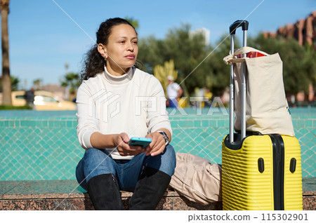 Woman sitting outdoors with luggage and cell phone, waiting for transport or enjoying a break Woman sitting outdoors with luggage and cell phone, waiting for transport or enjoying a break 115302901