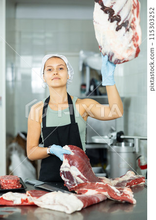 Girl in butcher shop cuts meat carcass into small pieces of meat for retail sale in small store 115303252