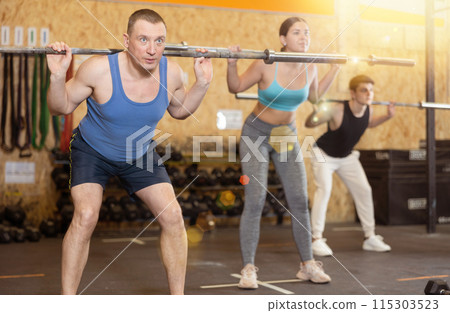 Focused middle-aged man in activewear exercising with barbell during group workout in gym health center. CrossFit healthy concept Focused middle-aged man in activewear exercising with barbell during group workout in gym health center. CrossFit healthy concept 115303523
