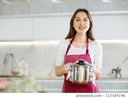 Smiling girl is standing in kitchen in dotted apron and holding stainless steel pot with lid Smiling girl is standing in kitchen in dotted apron and holding stainless steel pot with lid 115304146
