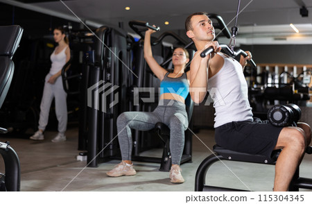 Slender athletic man work out muscles of arms and shoulders on simulator in gym of fitness club Slender athletic man work out muscles of arms and shoulders on simulator in gym of fitness club 115304345