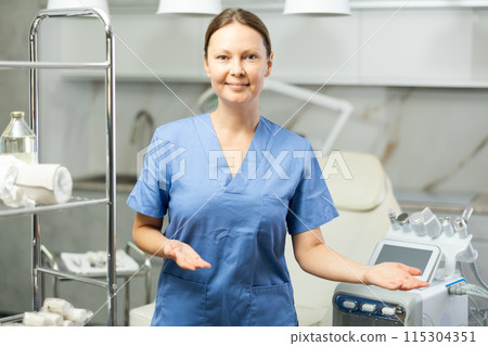 Portrait of middle-aged smiling masseuse doctor woman standing near beauty machine in medical salon, stretching hands aside with open palm, inviting clients 115304351
