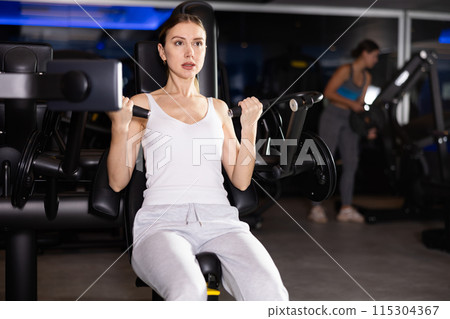 Sportive young girl training on arm curl machine in gym 115304367