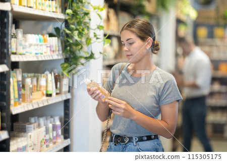 Positive young girl purchaser choosing cosmetic product in large supermarket 115305175