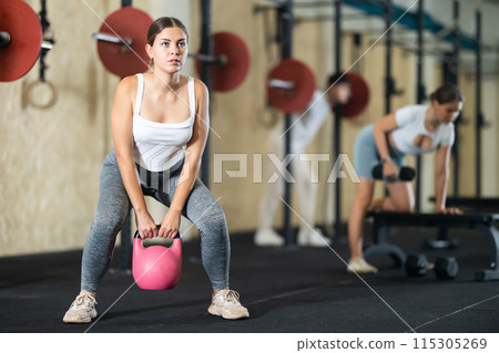 Young girl doing exercises with kettlebell near other people in crossfit gym 115305269