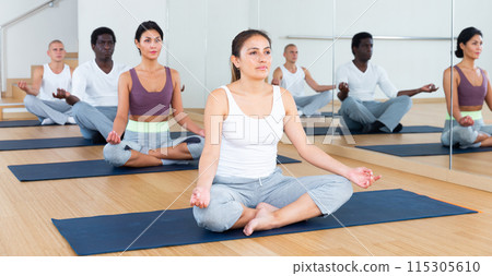 Young Latina meditating in lotus pose with group in fitness studio Young Latina meditating in lotus pose with group in fitness studio 115305610