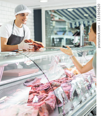 Positive young salesman demonstrating piece of meat to purchaser in butcher shop Positive young salesman demonstrating piece of meat to purchaser in butcher shop 115305620