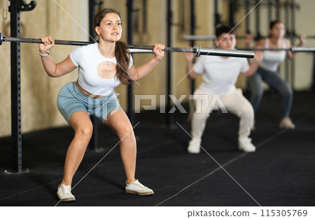 Girl in activewear exercising with barbell during group workout in gym center Girl in activewear exercising with barbell during group workout in gym center 115305769