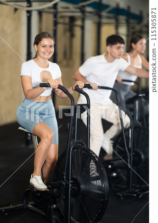 Positive girl riding exercise airbike for cardio workout during cycling class at modern crossfit gym 115305871