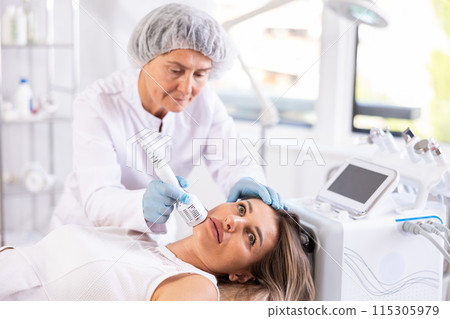 Female cosmetologist performing female face skin massage procedure in clinic Female cosmetologist performing female face skin massage procedure in clinic 115305979