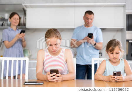 Sister girls sitting near kitchen table with phones in hands and flipping through tape Sister girls sitting near kitchen table with phones in hands and flipping through tape 115306032