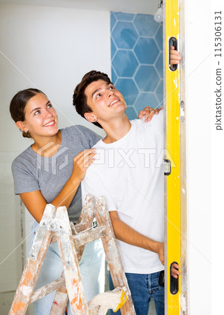 Positive couple checking laid tiles on wall with level meter in room under renovation Positive couple checking laid tiles on wall with level meter in room under renovation 115306131