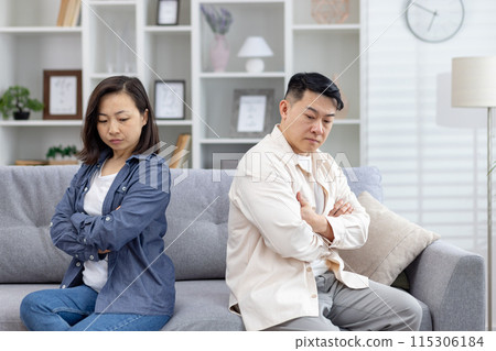 Asian couple feeling upset and distant while sitting on a couch in a living room. Emotions of conflict and disagreement are apparent between them. 115306184