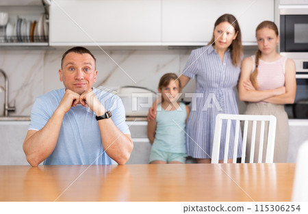 Upset father is sitting at the table, in background is a sad wife with small children. Family quarrel in kitchen Upset father is sitting at the table, in background is a sad wife with small children. Family quarrel in kitchen 115306254
