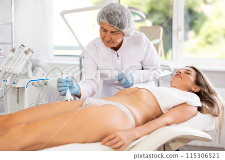 Mature female aesthetician administering high frequency ultrasoun procedure on woman thighs 115306521