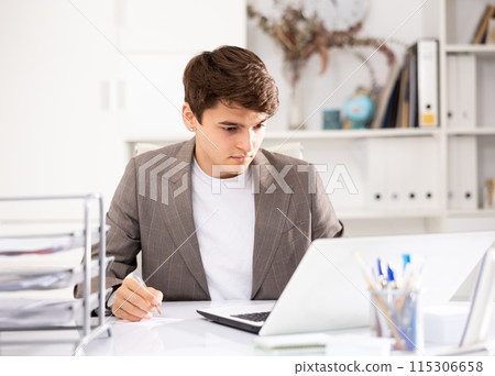 Young man office manager sitting at table and using laptop computer Young man office manager sitting at table and using laptop computer 115306658