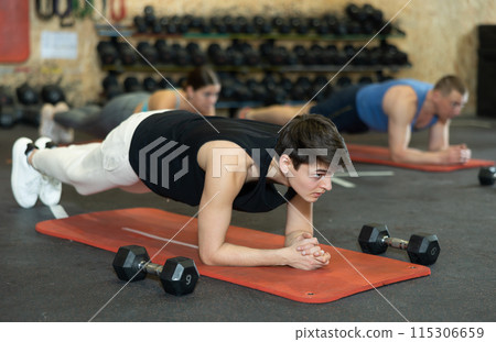 Energetic young man doing plank near other people in gym Energetic young man doing plank near other people in gym 115306659