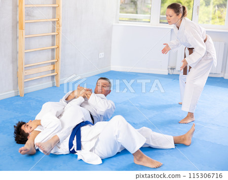 Judo or jiu-jitsu classes - two men practicing grabbing and throwing on sports mats under the guidance of trainer Judo or jiu-jitsu classes - two men practicing grabbing and throwing on sports mats under the guidance of trainer 115306716