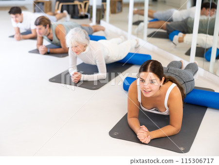 Young positive female performing set of exercises with pilates roller during group class in fitness studio. Young positive female performing set of exercises with pilates roller during group class in fitness studio. 115306717