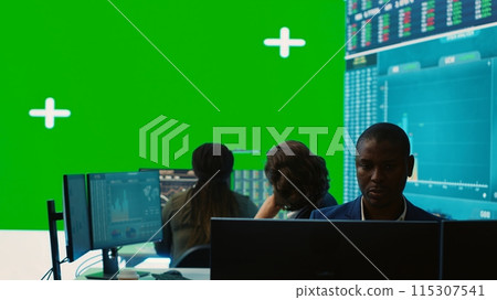 Diverse group of business specialists solve their daily tasks in control room with big greenscreen, analyzing big data numbers to ensure organizational progress. Startup management team. Camera A. 115307541