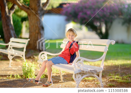 Cute blonde preteen tourist boy is sitting on a white bench in beautiful old park at summer sunset. Resort for family vacation. 115308586