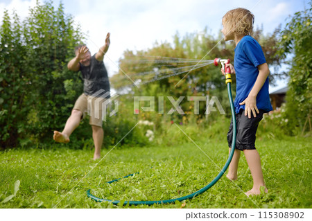 Funny little boy with his father playing with garden hose in sunny backyard. Preschooler child having fun with spray of water. Summer vacation in the village for family with kids. 115308902