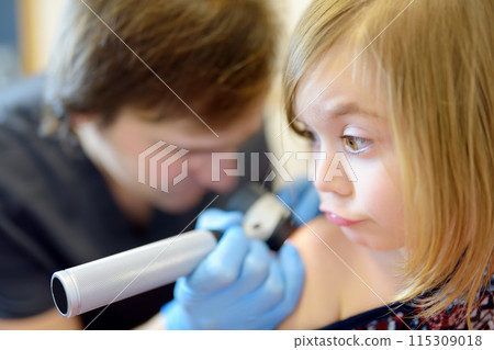 A caring doctor checks moles on the skin of a small child. A dermatologist looks at a rash on the back of a girl using a dermatoscope. 115309018
