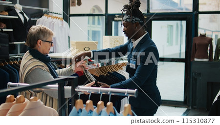 Elderly client asking to see multiple trendy bags in clothing store, talking to shop assistant about recommendations. African american worker shows different designer accessories. Camera B. 115310877