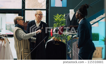 Store employee helping senior people with clothing suggestions, presenting new fashion collection items to couple in department store. African american employee makes recommendations. Camera A. Store employee helping senior people with clothing suggestions, presenting new fashion collection items to couple in department store. African american employee makes recommendations. Camera A. 115310881