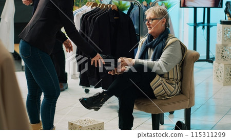 Retail worker presenting various pairs of shoes for senior client, assisting woman to find the perfect footwear in fashion showroom. Elderly customer trying on footgear before buying. Camera A. Retail worker presenting various pairs of shoes for senior client, assisting woman to find the perfect footwear in fashion showroom. Elderly customer trying on footgear before buying. Camera A. 115311299