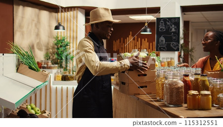 Local vendor placing crates with freshly harvested fruits on grocery store display, recommending organic produce to regular customer. African american farmer restocking all merchandise. Camera 2. 115312551