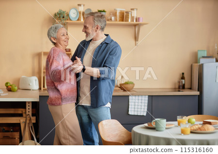 Side view portrait of happy senior couple slow dancing together in cozy kitchen and enjoying retirement copy space 115316160