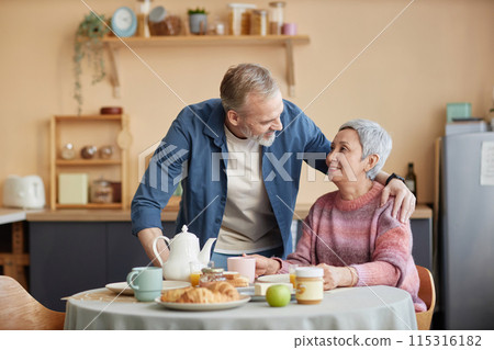 Front view portrait of affectionate senior couple enjoying breakfast together in cozy kitchen with husband embracing wife at table 115316182