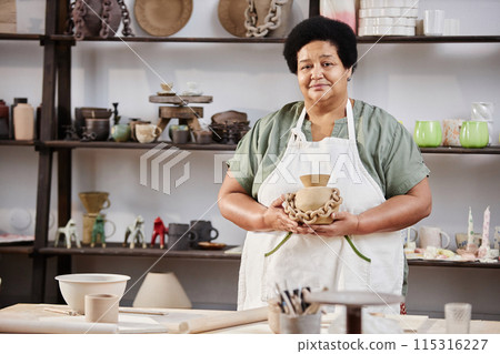 Waist up portrait of senior Black woman smiling at camera and holding handmade pottery piece in workshop, copy space Waist up portrait of senior Black woman smiling at camera and holding handmade pottery piece in workshop, copy space 115316227