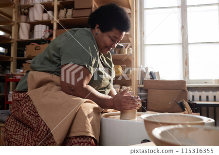 Side view portrait of smiling senior African American woman creating handmade ceramics on pottery wheel and enjoying cozy hobby in art studio copy space Side view portrait of smiling senior African American woman creating handmade ceramics on pottery wheel and enjoying cozy hobby in art studio copy space 115316355