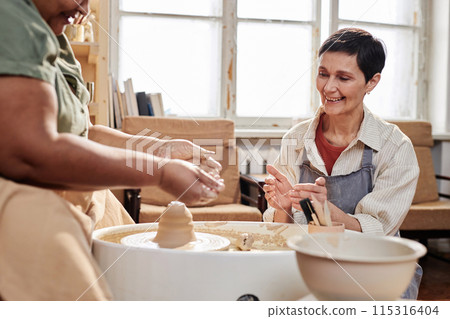 Portrait of smiling mature woman as art teacher assisting student creating ceramics in cozy studio copy space Portrait of smiling mature woman as art teacher assisting student creating ceramics in cozy studio copy space 115316404