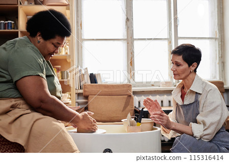 Side view portrait of Black senior woman enjoying pottery class in sunlight with female art teacher assisting Side view portrait of Black senior woman enjoying pottery class in sunlight with female art teacher assisting 115316414