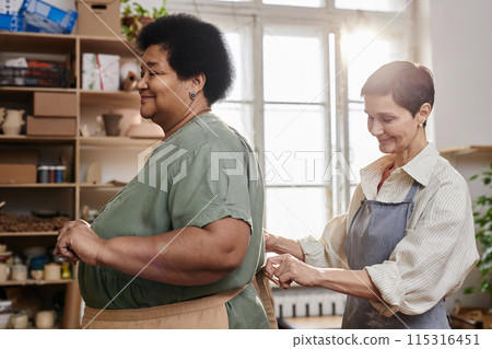 Side view portrait of two smiling adult women tying aprons preparing for art class in creative studio in sunlight 115316451