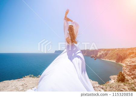 woman sea white dress. Blonde with long hair on a sunny seashore in a white flowing dress, rear view, silk fabric waving in the wind. Against the backdrop of the blue sky and mountains on the seashore woman sea white dress. Blonde with long hair on a sunny seashore in a white flowing dress, rear view, silk fabric waving in the wind. Against the backdrop of the blue sky and mountains on the seashore 115320858