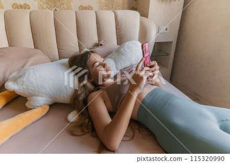 A girl is laying on a bed with a pink phone in her hand. She is looking at the phone and she is focused on something on the screen. A girl is laying on a bed with a pink phone in her hand. She is looking at the phone and she is focused on something on the screen. 115320990
