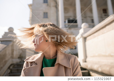 A woman with long hair is standing on a set of stairs. She is wearing a green shirt and a tan coat. 115321007