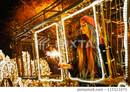 Woman holding sparkler night while celebrating Christmas outside. Dressed in a fur coat and a red headband. Blurred christmas decorations in the background. Selective focus 115321073