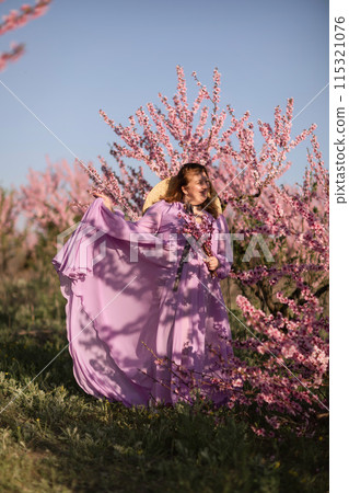 Woman blooming peach orchard. Against the backdrop of a picturesque peach orchard, a woman in a long pink dress and hat enjoys a peaceful walk in the park, surrounded by the beauty of nature. 115321076