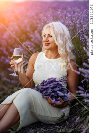 Blonde lavender field holds a glass of white wine in her hands. Happy woman in white dress enjoys lavender field picnic holding a large bouquet of lavender in her hands . Illustrating woman's picnic 115321148