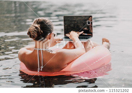 A woman is floating on a pink inflatable raft in the ocean while using a laptop. Concept of relaxation and leisure, as the woman enjoys her time in the water while working on her laptop. A woman is floating on a pink inflatable raft in the ocean while using a laptop. Concept of relaxation and leisure, as the woman enjoys her time in the water while working on her laptop. 115321202