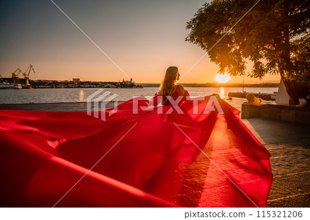 Sunrise red dress. A woman in a long red dress against the backdrop of sunrise, bright golden light of the sun's rays. The concept of femininity, harmony. 115321206