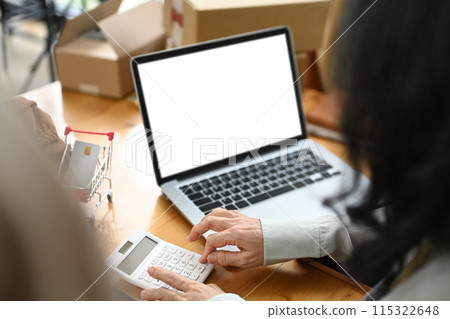 Small business owner sitting in front of laptop with blank screen and calculating expenses Small business owner sitting in front of laptop with blank screen and calculating expenses 115322648