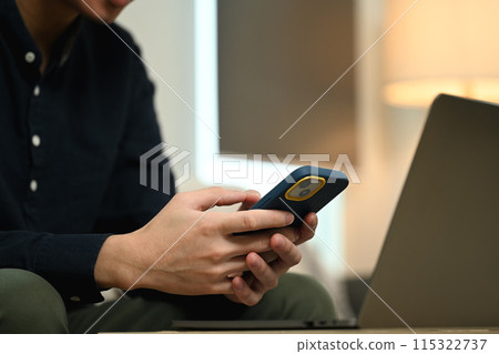 Male freelancer working with laptop and using mobile phone on couch in living room 115322737